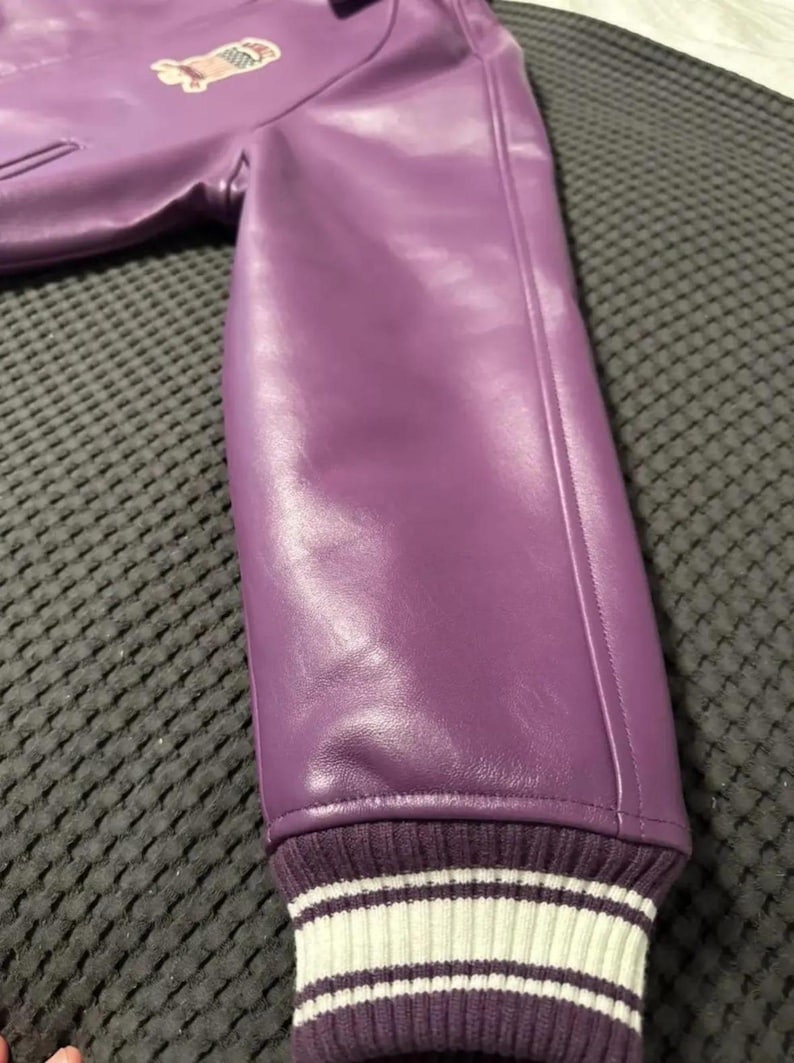 May include: A close-up of a purple leather jacket sleeve. The sleeve has a ribbed cuff with white stripes. A small patch is visible on the upper part of the jacket. The jacket is made of a smooth, shiny material.