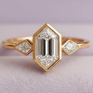Puede incluir: Un anillo de compromiso de oro con una gran piedra preciosa rectangular y transparente en el centro, flanqueada por dos piedras más pequeñas en forma de diamante. El anillo está engastado en una banda de oro y se muestra en un joyero morado.