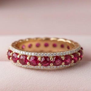 Pode incluir: Um anel de ouro com uma faixa de pedras preciosas vermelhas vibrantes, circundado por uma fileira de pedras menores e transparentes. O anel é apresentado numa caixa de joias de veludo rosa, criando uma exibição luxuosa.