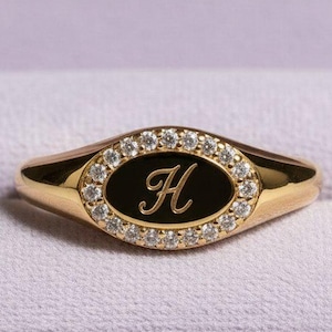 May include: A gold signet ring featuring an oval-shaped black inlay centred with a script letter H. The oval inlay is surrounded by a halo of small, clear, round-cut stones set into the gold band, creating a polished and reflective appearance.