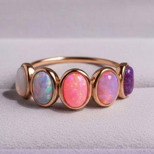 May include: A rose gold ring featuring five oval-shaped opals in shades of white, blue, pink, and purple. The ring is set in a delicate band and is displayed inside a purple jewelry box.