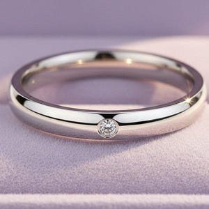 May include: A silver ring with a single, small, round diamond set into the band. The ring is displayed in a soft purple ring box, with the focus on the ring's polished surface and the sparkle of the diamond.