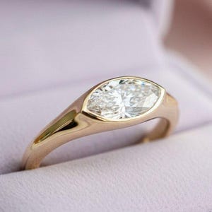 May include: A gold ring featuring a marquise-cut diamond, set within a bezel. The ring is presented in a light purple box. The polished gold band curves smoothly. The diamond's facets catch the light, creating a brilliant sparkle.