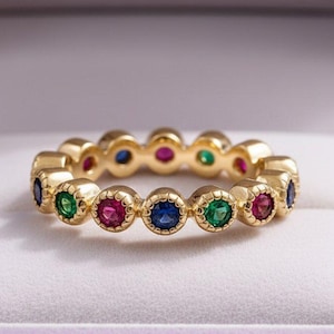 May include: A gold band ring featuring a series of round, bezel-set gemstones in alternating colors of vibrant blue, bright green, and deep pink. The metal setting around each individual stone has a textured, scalloped edge detail.