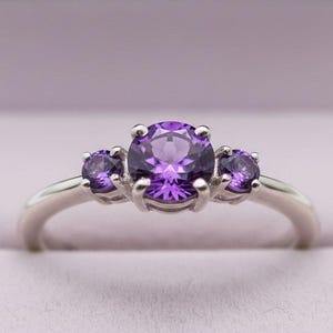 May include: A silver ring featuring three round, vibrant purple gemstones. The central stone is larger, flanked by two smaller stones. The ring is set against a soft purple background, suggesting a jewelry box.