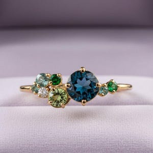 Può includere: Un anello d'oro con un gruppo di pietre preziose. L'anello presenta una grande pietra centrale rotonda di colore verde-azzurro scuro, circondata da pietre verdi, verde-azzurro e bianche più piccole. L'anello è incastonato in una fascia d'oro e viene presentato in un cofanetto viola.