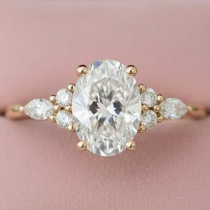 May include: A gold engagement ring with an oval-cut diamond center stone. The ring features smaller round and pear-shaped diamonds on the band. The ring is set in a pink jewelry box.