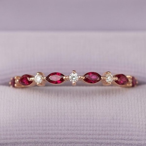 May include: A thin gold band ring featuring an alternating pattern of marquise-cut deep red rubies and small round brilliant-cut white diamonds. The stones are set in a delicate, symmetrical arrangement along the center of the metal band.
