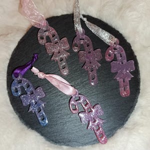 Può includere: Ornamenti a forma di bastoncino di zucchero glitterati nei toni del rosa, viola e argento, ciascuno con un fiocco. Gli ornamenti sono sospesi da nastri di vari colori. Sono disposti su una superficie circolare grigio scuro.