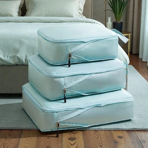 Pode incluir: Três cubos de armazenamento azul claro empilhados. Cada cubo tem um fecho de zíper com um puxador preto e uma alça de tecido. Os cubos são feitos de um material texturizado, ideais para organizar itens.