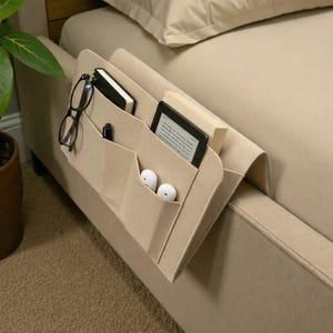 Puede incluir: Organizador de fieltro beige para mesita de noche con múltiples bolsillos. El organizador está colocado sobre el brazo de un sofá beige y contiene un cuaderno, gafas, bolígrafo, lector electrónico y auriculares. Está diseñado para tener lo esencial a mano.