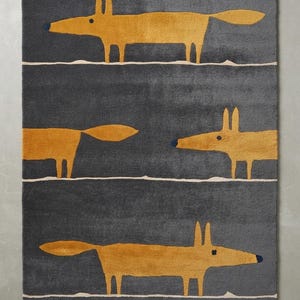 Peut inclure: Un tapis rectangulaire au fond gris foncé, divisé en trois rangées horizontales par de fines lignes claires. Chaque rangée présente une figure de renard orange stylisée avec un museau bleu foncé, orientée dans des directions alternées.