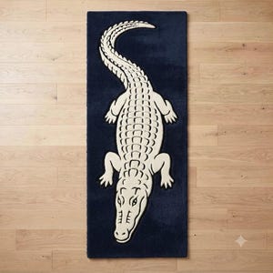 Könnte beinhalten: Ein rechteckiger Teppich mit dunkelblauem Hintergrund und einer großen, cremefarbenen Alligatorgrafik in der Mitte. Der Alligator ist aus der Vogelperspektive dargestellt, wobei der Schwanz nach oben gebogen ist und der Kopf nach unten zeigt.