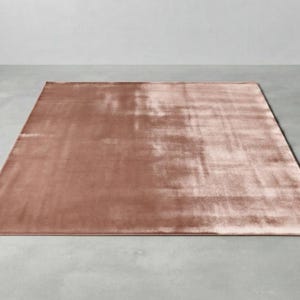 Peut inclure: Un tapis rectangulaire d'une couleur rose poudré unie. La surface présente une texture lisse semblable à du velours avec de subtiles variations de réflexion de la lumière sur le tissu, créant un éclat doux. Une petite étiquette noire est visible.
