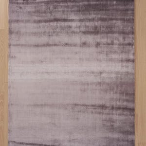 Op de afbeelding: Een rechthoekig vloerkleed in grijstinten en paars. Het vloerkleed heeft een zachte, fluweelachtige textuur met een subtiel horizontaal streepjespatroon. Het vloerkleed ligt op een lichtgekleurde houten vloer.