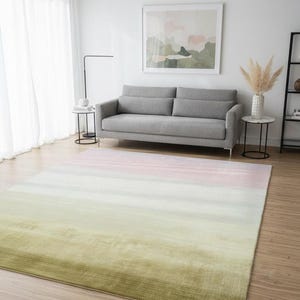 Pode incluir: Um tapete retangular grande com um gradiente de cores, que vai do verde ao branco e ao rosa. O tapete está colocado sobre um chão de madeira numa sala de estar, com um sofá cinzento e obras de arte no fundo. As dimensões do tapete são em centímetros.