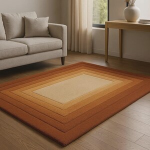 Könnte beinhalten: Ein rechteckiger Teppich mit einem mehrschichtigen Design in Beige-, Hellbraun- und Rosttönen. Der Teppich hat ein zentrales beiges Rechteck, das von progressiv dunkleren Rändern umgeben ist und einen Farbverlauf erzeugt. Der Teppich liegt auf einem Holzboden.