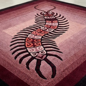 Puede incluir: Una alfombra rectangular con un gráfico grande y estilizado de un ciempiés en tonos rojo, rosa y burdeos. El ciempiés tiene numerosas patas oscuras y un cuerpo segmentado con patrones geométricos. Incluye una pequeña etiqueta marrón con texto en hindi.