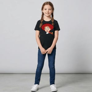 May include: A young girl wearing a black t-shirt featuring a cartoon image of a character with red hair and a red hat. She is also wearing blue jeans and white sneakers. The girl is standing in front of a gray background.