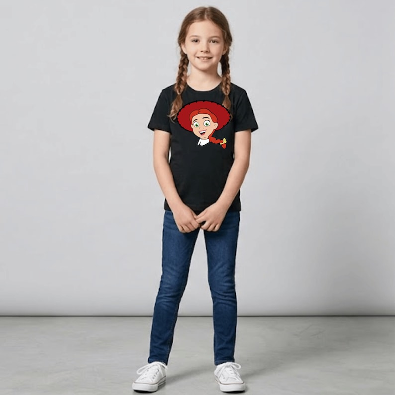 May include: A young girl wearing a black t-shirt featuring a cartoon image of a character with red hair and a red hat. She is also wearing blue jeans and white sneakers. The girl is standing in front of a gray background.