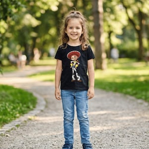 May include: A young girl wearing a black t-shirt featuring a cartoon image of Jessie from Toy Story. The shirt is paired with blue jeans and blue shoes. The girl is smiling and standing on a gravel path in a park setting.