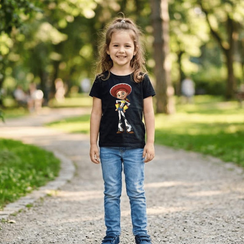 May include: A young girl wearing a black t-shirt featuring a cartoon image of Jessie from Toy Story. The shirt is paired with blue jeans and blue shoes. The girl is smiling and standing on a gravel path in a park setting.