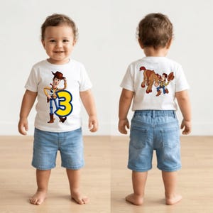 May include: A toddler wearing a white t-shirt with a cartoon image of Woody from Toy Story and the number 3 in yellow and blue. The toddler is also wearing blue denim shorts.