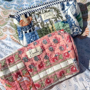 May include: Two quilted cosmetic bags. The top bag has a patchwork design with blue, green, and white floral patterns. The bottom bag is pink with a floral print and white stripes. Both bags have a zip closure and a label that says "Brimela".