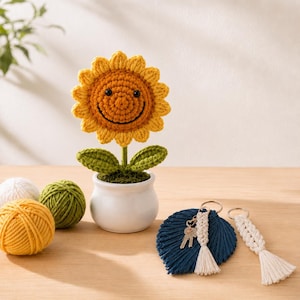 Può includere: Un girasole all'uncinetto con un viso sorridente in un vaso di ceramica bianca, accompagnato da due gomitoli di lana gialla e verde, e due portachiavi in macramè con una forma a foglia blu scuro e un cordino intrecciato bianco con nappa.