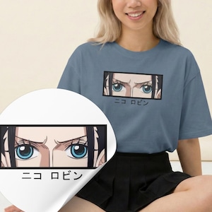 May include: A blue t-shirt featuring a rectangular graphic of anime eyes with blue irises and the Japanese text "ニコ ロビン". The graphic is framed in black. The shirt is a solid blue color.
