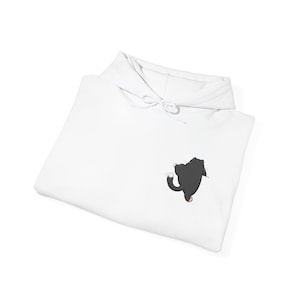 Puede incluir: Sudadera con capucha blanca con cordón. En la parte delantera hay una pequeña ilustración de dibujos animados de un gato negro con la cola con punta blanca. El gato está en una pose juguetona. La sudadera con capucha está doblada.