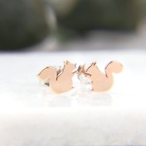 May include: A pair of rose gold stud earrings shaped like squirrels. Each earring features a smooth, polished finish and a silhouette of a squirrel with a curled tail, designed with a post back for secure attachment to the earlobe.