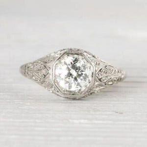 Puede incluir: Un anillo detallado de oro blanco con un engaste octogonal y una gran gema transparente. El anillo presenta intrincados diseños de filigrana a lo largo de la banda y alrededor de la piedra central, mostrando una estética vintage.