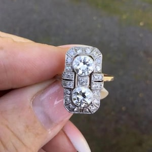 Puede incluir: Anillo de estilo Art Déco con dos grandes piedras redondas y transparentes. El engaste rectangular, de color plateado, está adornado con piedras claras más pequeñas. La banda del anillo es dorada.