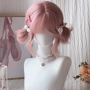 Puede incluir: Una peluca rosa pastel con flequillo y coletas, sujeta con accesorios para el cabello rosas y blancos. La peluca se exhibe en una cabeza de maniquí blanca, complementada con un collar de encaje con un colgante en forma de corazón. Una pintura de zapatillas de ballet está en el fondo.