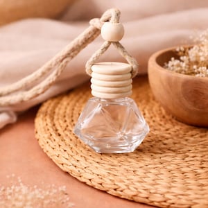 May include: A clear, faceted glass car air freshener bottle with a wooden cap and a beige rope for hanging. The bottle sits on a woven straw mat, with a wooden bowl and dried flowers in the background. The overall colour palette is neutral.