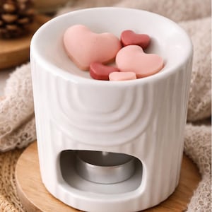 Puede incluir: Un calentador de cera de cerámica blanca con una base cilíndrica y una parte superior en forma de plato. El plato contiene ceras perfumadas en forma de corazón en tonos rosa y rojo. Una pequeña vela de té está dentro.
