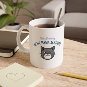 Puede incluir: Taza de cerámica blanca con el texto "Me Looking at My Bank Account" y una imagen de dibujos animados de un gato gris enfadado. La taza contiene un líquido oscuro y una cuchara plateada.