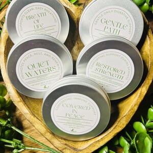 May include: Five small, silver tins with white labels are arranged in a wooden heart-shaped bowl, surrounded by greenery. Each tin features text like "Breath of Life" and "Quiet Waters". The labels also list ingredients and Bible verses.