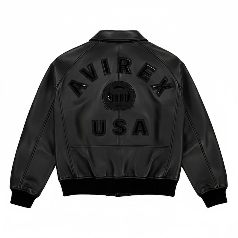 Puede incluir: Chaqueta bomber de cuero negro con la palabra "AVIREX" arqueada en la espalda, encima de un emblema circular y la palabra "USA". La chaqueta tiene cuello, pu&ntilde;os y dobladillo de canal&eacute;.