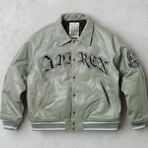 May include: A light green leather jacket with the text "AVI REX" in black script across the chest. The jacket has a button-up front, side pockets, and a ribbed hem and cuffs with white stripes. A patch is on the upper left sleeve.