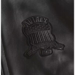 Puede incluir: Primer plano de una chaqueta de cuero negro con una superficie texturizada. Un parche negro con el texto "AVIREX" y "ESTABLISHED 1975" est&aacute; cosido en la chaqueta. El parche tiene un dise&ntilde;o de escudo.