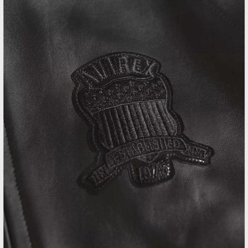 Puede incluir: Primer plano de una chaqueta de cuero negro con una superficie texturizada. Un parche negro con el texto "AVIREX" y "ESTABLISHED 1975" est&aacute; cosido en la chaqueta. El parche tiene un dise&ntilde;o de escudo.