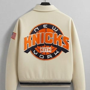 May include: Cream-colored jacket with a black collar and striped cuffs. The back features a large orange and navy graphic of a basketball with the words "New York Knicks" and "KITH" in a circular design. An American flag patch is on the left sleeve.