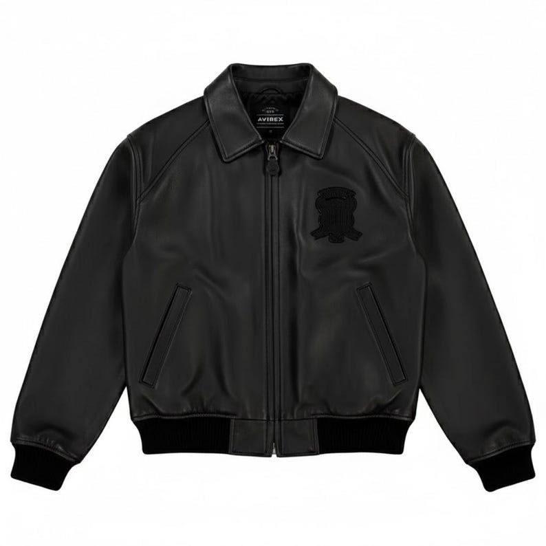 Puede incluir: Chaqueta de cuero negra con cuello, cierre de cremallera y un emblema negro en el pecho. La chaqueta tiene dos bolsillos delanteros y pu&ntilde;os y dobladillo acanalados. La marca "AVIREX" es visible en el cuello.