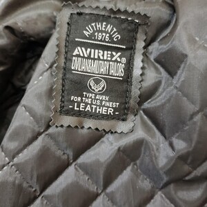Puede incluir: Primer plano de una etiqueta de chaqueta de cuero negra. La etiqueta dice "AUTHENTIC 1975 AVIREX CIVILIAN & MILITARY TAILORS". Debajo del logotipo, dice "TYPE AVRX FOR THE U.S. FINEST - LEATHER -."