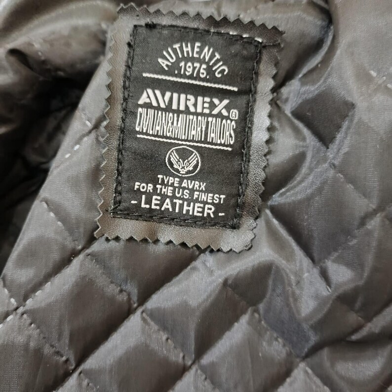 Puede incluir: Primer plano de una etiqueta de chaqueta de cuero negra. La etiqueta dice "AUTHENTIC 1975 AVIREX CIVILIAN & MILITARY TAILORS". Debajo del logotipo, dice "TYPE AVRX FOR THE U.S. FINEST - LEATHER -."
