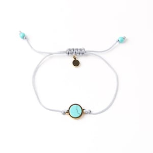 May include: A delicate bracelet with a round, light blue stone in a gold-coloured bezel. The bracelet has a grey cord with a sliding knot closure and small turquoise beads at the ends. A small gold charm hangs near the knot.