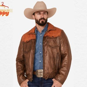 May include: A brown leather jacket with a contrasting orange yoke and collar. The jacket has two chest pockets and a button closure. The model is wearing a cowboy hat and a denim shirt. The belt buckle is silver and gold.