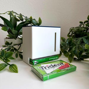 May include: A white rectangular plastic dispenser with a dark blue top edge and a vertical slot on the front. A pack of green Trident Watermelon Twist gum is inserted into the bottom opening, and another pack of the same gum lies in front.